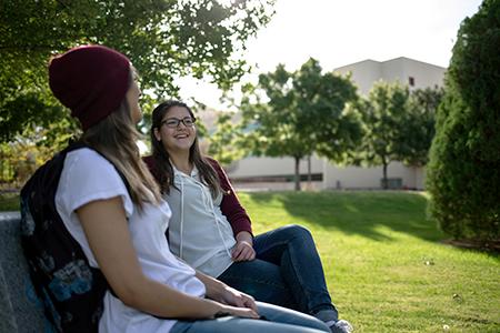 两个女学生坐在长凳上聊天的画面. 在图像背景中可以看到一座建筑物的剪影.